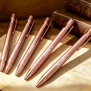 Puede incluir: Cinco bolígrafos de oro rosa con nombres personalizados: James, Emily, Amelia, Olivia y Charlotte. Cada bolígrafo tiene un diseño elegante con punta táctil y clip para bolsillo. Se exhiben sobre papel antiguo, con libros antiguos al fondo.