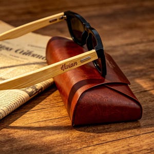 Pu&ograve; includere: Occhiali da sole con montatura nera e aste in legno chiaro, appoggiati su un astuccio in pelle marrone. Il nome "Vivian" &egrave; stampato sull'asta degli occhiali. Gli occhiali sono su un giornale.