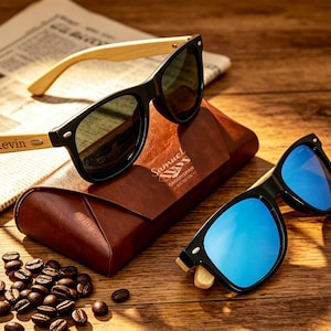 Pu&ograve; includere: Due paia di occhiali da sole con montatura nera e aste in legno sono appoggiati su un astuccio in pelle marrone. Un paio ha lenti verdi, l'altro lenti blu. La parola "Kevin" &egrave; incisa su un'asta. Chicchi di caff&egrave; e un giornale sullo sfondo.