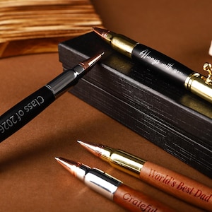 Pode incluir: Uma coleção de canetas projetadas para se assemelharem a balas. As canetas apresentam detalhes metálicos e são gravadas com frases como "Always with you", "Class of 2026", "World's Best Dad" e "Grateful". As canetas são exibidas em uma superfície marrom.