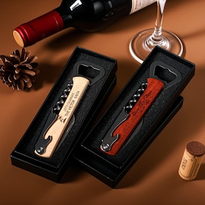 Puede incluir: Dos juegos de abridor de botellas y sacacorchos personalizados en cajas negras. Uno es de madera clara con grabado, el otro de madera oscura con grabado. Una botella de vino y una copa están en el fondo.