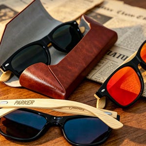 Pu&ograve; includere: Occhiali da sole con montatura nera e aste in legno sono esposti con un astuccio in pelle marrone. Un paio ha lenti blu, un altro lenti rosse e un terzo paio &egrave; nell'astuccio. La parola "PARKER" &egrave; incisa sull'asta di un paio.
