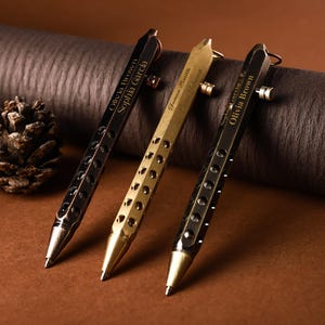 Op de afbeelding: Drie metalen pennen in zwart, goud en brons, elk met een uniek ontwerp en gepersonaliseerde tekst. De pennen hebben een zeshoekige schacht met cirkelvormige uitsparingen en een punt. De tekst bevat namen als Olivia Brown en Sophia Garcia.