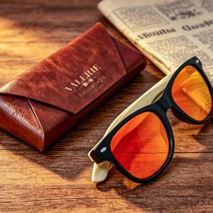 Pu&ograve; includere: Occhiali da sole con montatura nera e lenti arancioni su una superficie di legno, accanto a un astuccio in pelle marrone. L'astuccio riporta la scritta "VALERIE GROOMSMEN". Un giornale &egrave; sullo sfondo.
