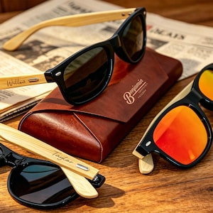 Pu&ograve; includere: Occhiali da sole con montatura nera e aste in bamb&ugrave; sono esposti su una superficie di legno. Un paio ha lenti a specchio arancioni. &Egrave; presente anche un astuccio in pelle marrone con il nome "Benjamin". Le aste degli occhiali sono incise con nomi.