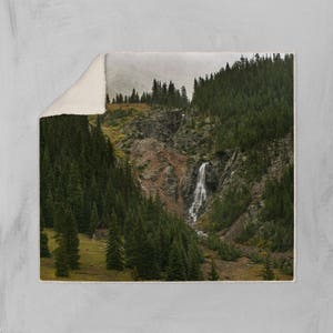 May include: A scenic landscape photograph featuring a waterfall cascading down a rocky mountainside, surrounded by lush green trees. A small wooden cabin is nestled in the trees at the base of the mountain.
