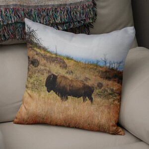May include: A square throw pillow featuring a photographic print of a brown American bison standing in a field of dry, golden-brown grass on a hillside. The scene includes rocky terrain, sparse bare trees, and a pale, overcast sky in the background.