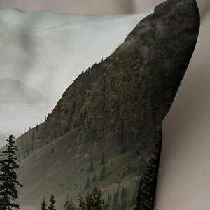 May include: A close-up of a mountainside with a rocky, green, and brown texture. The mountain is covered in a light mist, and there are trees growing on the slopes.