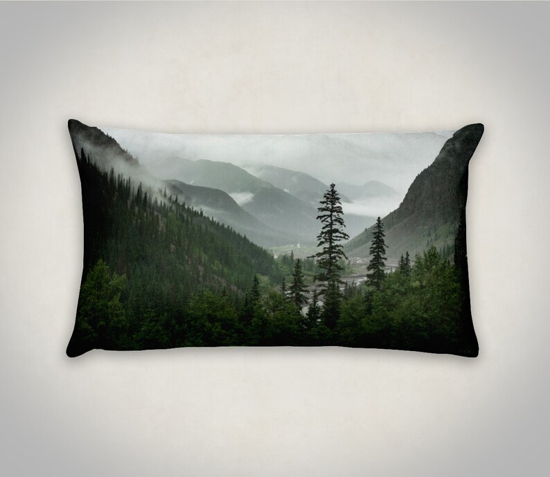 May include: A scenic pillow featuring a misty mountain landscape with a tall evergreen tree in the foreground. The mountains are covered in lush green trees and fog.
