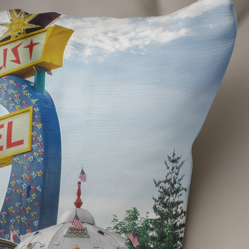 May include: A pillow with a vintage motel sign featuring a blue and yellow star with the word "Star" on it. The sign is partially obscured by a white cloud in a blue sky. The pillow also features a white building with a red and white striped dome and American flags.
