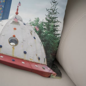 May include: A white and red dome-shaped structure with a round window and blue and red accents. The structure is topped with a spire and two American flags. The structure is surrounded by green trees.