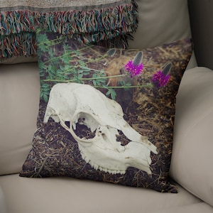 Könnte beinhalten: Ein weißer Tierschädel mit Zähnen liegt auf einem Bett aus braunen Kiefernnadeln und grünem Laub. Im Hintergrund befinden sich violette Wildblumen.