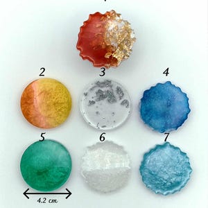 Peut inclure: Collection de sept pièces d'art en résine de différentes couleurs et motifs. Les pièces rondes mesurent 4,2 cm de diamètre et 0,8 cm d'épaisseur. Certaines ont des bords texturés et des accents métalliques. L'épaisseur avec prise est de 1,4 cm.