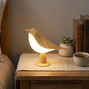 Peut inclure: Lampe de table en forme d'oiseau en bois, dégageant une lumière chaleureuse. Le corps de la lampe est en bois brun clair, avec un ventre blanc illuminé, posée sur un socle en bois. Sur une table de chevet en bois.