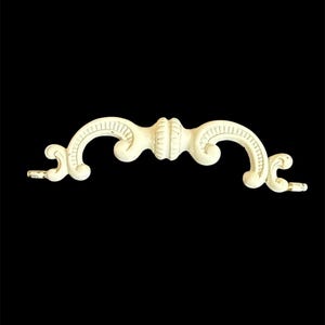 May include: Off-white, ornate furniture handle with a symmetrical design. The handle features curved ends and a central, decorative element. The piece is set against a black background, highlighting its intricate details and vintage aesthetic.