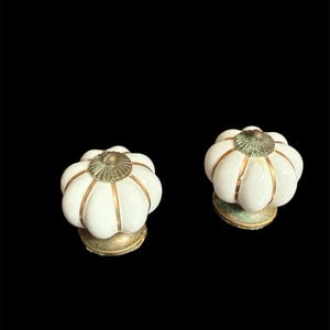 May include: Two white ceramic cabinet knobs with a pumpkin-like design. Each knob features gold-colored accents and a brass-colored base. The knobs are set against a black background.
