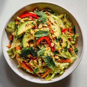 May include: A vibrant salad in a white bowl, featuring sliced avocado, red bell peppers, shredded carrots, and fresh mint leaves. The salad is topped with peanuts and a creamy dressing, creating a colorful and appetizing dish.