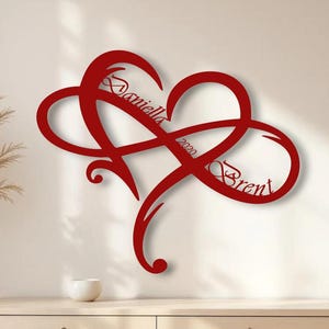 Può includere: Arte da parete in metallo rosso raffigurante un cuore intrecciato con un simbolo dell'infinito. I nomi "Daniella" e "Brent" sono incisi, insieme all'anno "2020". L'opera è montata su una parete neutra sopra un comò in legno.