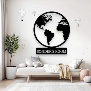May include: Black metal wall art depicting a globe with the text "SONDER'S ROOM" underneath. The artwork is mounted on a white wall, decorated with hot air balloon silhouettes. A bed, pillows, and a potted plant are in the foreground.