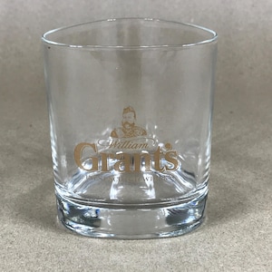May include: A clear, tumbler-style glass with a thick base. The glass has a gold-coloured "William Grant's" logo, including an illustration of a man in a kilt. The words "FINEST SCOTCH WHISKY" are printed below the logo. The glass is empty.