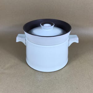 May include: A white ceramic cooking pot with a dark brown interior and lid. The pot has two side handles and a small knob on the lid. The pot is designed for cooking and serving food.