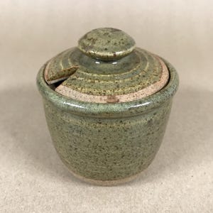 May include: A handmade, speckled green ceramic sugar bowl with a lid and a small knob. The lid has a slot for a spoon. The bowl is a muted green colour with a light brown band around the middle.