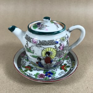 May include: A small, white ceramic teapot with a matching saucer. The teapot and saucer are decorated with a colourful Asian-inspired scene featuring figures, buildings, and floral elements. The teapot has a green rim and handle.