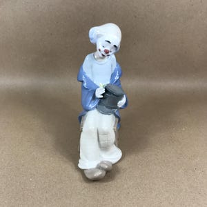 May include: A porcelain clown figurine. The clown has a white head and face, red lips, and blue eye shadow. It wears a light blue shirt, a blue jacket, and white trousers. The clown holds a grey top hat.