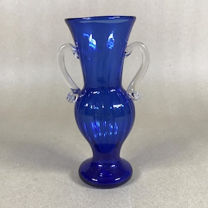 May include: A vibrant cobalt blue glass vase featuring two clear glass handles. The vase has a flared top and a rounded base. Vertical ridges add texture to the body. A decorative item for home interiors.