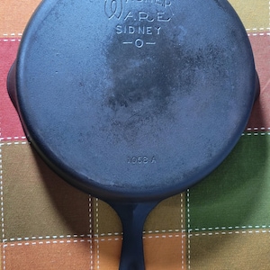 May include: A round black cast iron skillet with a long handle. The bottom features embossed text reading WAGNER WARE SIDNEY -O- and the number 1058 A. The surface shows a dark, matte finish with subtle variations in color and texture.