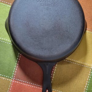 May include: A black cast iron skillet with a long handle and a circular cooking surface. The skillet has the text "Wagner Ware Sidney -0-" and the number "1058W" embossed on the bottom. The skillet is on a colorful checkered surface.