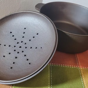 May include: A dark brown, oval-shaped cast iron Dutch oven with a matching lid. The lid has a pattern of small holes in a starburst design. The oven has two handles and sits on a colorful, patterned surface.