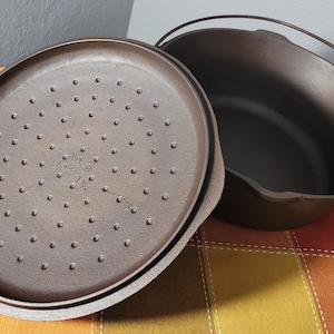 May include: A cast iron Dutch oven with a lid. The lid is perforated with small holes and has the text "NO. 8 10 IN." The pot has a handle and a dark brown finish. The pot and lid are on a colorful checkered surface.