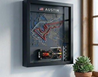 May include: A black framed wall art piece featuring a layered 3D map of the Austin race track with a red circuit line, a small American flag icon, the text AUSTIN, and a miniature Formula 1 race car model mounted at the bottom with a finish line.