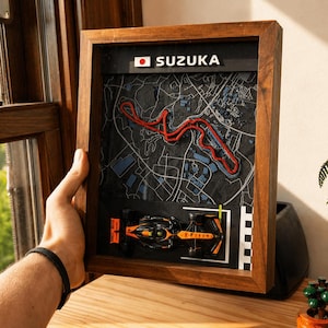 May include: A framed 3D wall art piece featuring a map of the Suzuka race track in Japan. The display includes a red track outline on a dark gray background, a Japanese flag icon, the text SUZUKA, and a miniature black and orange race car model.