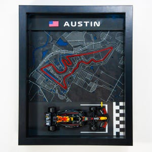 May include: Framed artwork featuring a map of the Austin, Texas race track with a miniature race car. The map is in black, white, red, and blue, with the word "AUSTIN" and the American flag at the top. The race car is black, red, and yellow.