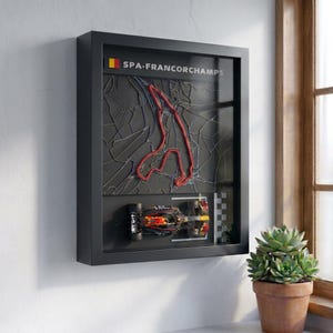 Puede incluir: Un cuadro de pared con marco negro que presenta un mapa topográfico en 3D del circuito de Spa-Francorchamps con una línea roja. Debajo del mapa, un coche de carreras de Fórmula 1 en miniatura se sitúa junto a una línea de meta a cuadros.