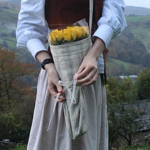 May include: A fabric flower holder with a plaid pattern, filled with yellow roses. The holder is secured with a white ribbon. The person is wearing a white blouse, brown vest, and long skirt.