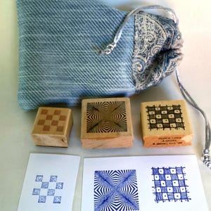 May include: A set of three wooden stamps with geometric designs, including a checkerboard and radial patterns. The stamps are accompanied by printed examples in blue ink on white paper. A blue drawstring bag with a paisley pattern holds the set.