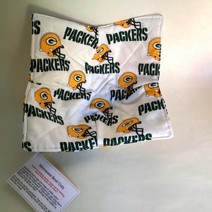 May include: White microwave bowl cozy with a repeating pattern of green and yellow football helmets and the word "Packers". The cozy is designed to keep bowls cool to the touch after microwaving. The item is made of 100% cotton.