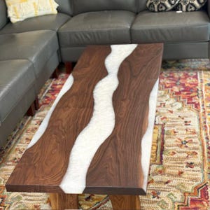 Peut inclure: Une table basse rectangulaire avec une surface en bois brun foncé et un motif blanc en forme de rivière traversant le centre. La table repose sur des pieds en bois et est posée sur un tapis coloré.