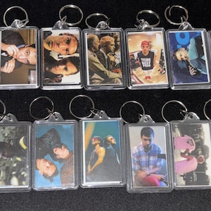 Puede incluir: Una colección de llaveros rectangulares con varias imágenes en su interior. Los llaveros tienen un marco de plástico transparente y un anillo de metal plateado. Las imágenes parecen ser de personas, algunas en color y otras en blanco y negro.