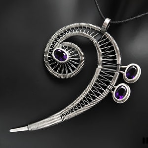 Può includere: Una collana con pendente argentato a forma di chiave di violino, con tre pietre preziose viola di forma ovale. Il pendente è finemente lavorato a filo e sospeso a un cordino nero. Lo sfondo è nero.