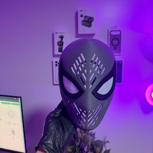 May include: A dark gray, textured full-face mask featuring a stylized, angular web-like pattern of cutouts across the forehead and mouth area, complemented by large, almond-shaped eye openings with black borders and white mesh lenses.