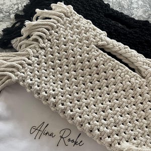 May include: Close-up of macrame items in cream and black. The cream-coloured macrame has a woven pattern with fringe. A white cloth with the text "Alina Rooke" is visible. The black macrame has a similar woven pattern and fringe.