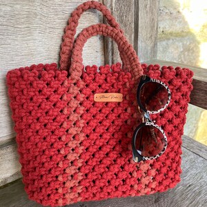 May include: A red macrame tote bag with matching handles. The bag features a woven design and a small leather tag. Sunglasses with a black and white pattern are attached.