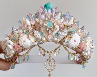 Puede incluir: Una corona blanca y dorada adornada con conchas marinas, perlas y cristales. La corona tiene un encanto de luna creciente y estrella colgando de la parte inferior.