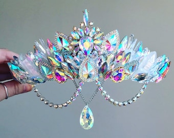 May include: A silver crown featuring iridescent crystals and a teardrop pendant. The crown is adorned with white feathers and crystals.