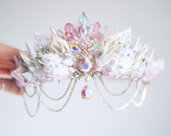 May include: A white and pink crown with seashells, crystals, and a chain of pearls. The crown is decorated with iridescent rhinestones and has a dangling crystal pendant.