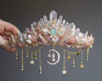 Puede incluir: Una corona delicada hecha de cristales de cuarzo transparentes, conchas marinas y detalles dorados. La corona presenta un dije de luna creciente con estrellas y cadenas colgantes con dijes de estrellas.